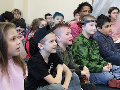 A group of young special education students sit together on the floor while listening to a presentation.