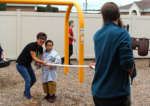 Special education teachers on a playground teach a young boy to play tug of war.