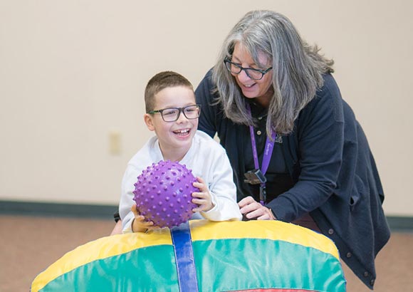 Staff member helping happy special education boy practice his balance by rolling on top of the rocking barrel to retrieve a ball in an occupational therapy session.
