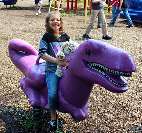 A young girl holds her stuffed animal while riding a purple dinosaur playground toy