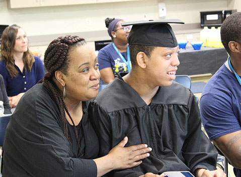 A mother smiles with her son in his cap and gown as he waits to graduate