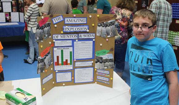 A young boy stands beside his science fair project at his special education school