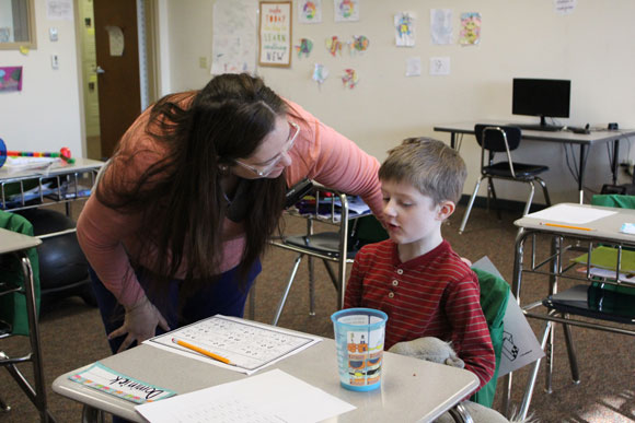A special education teacher leans over an elementary school boy during class.