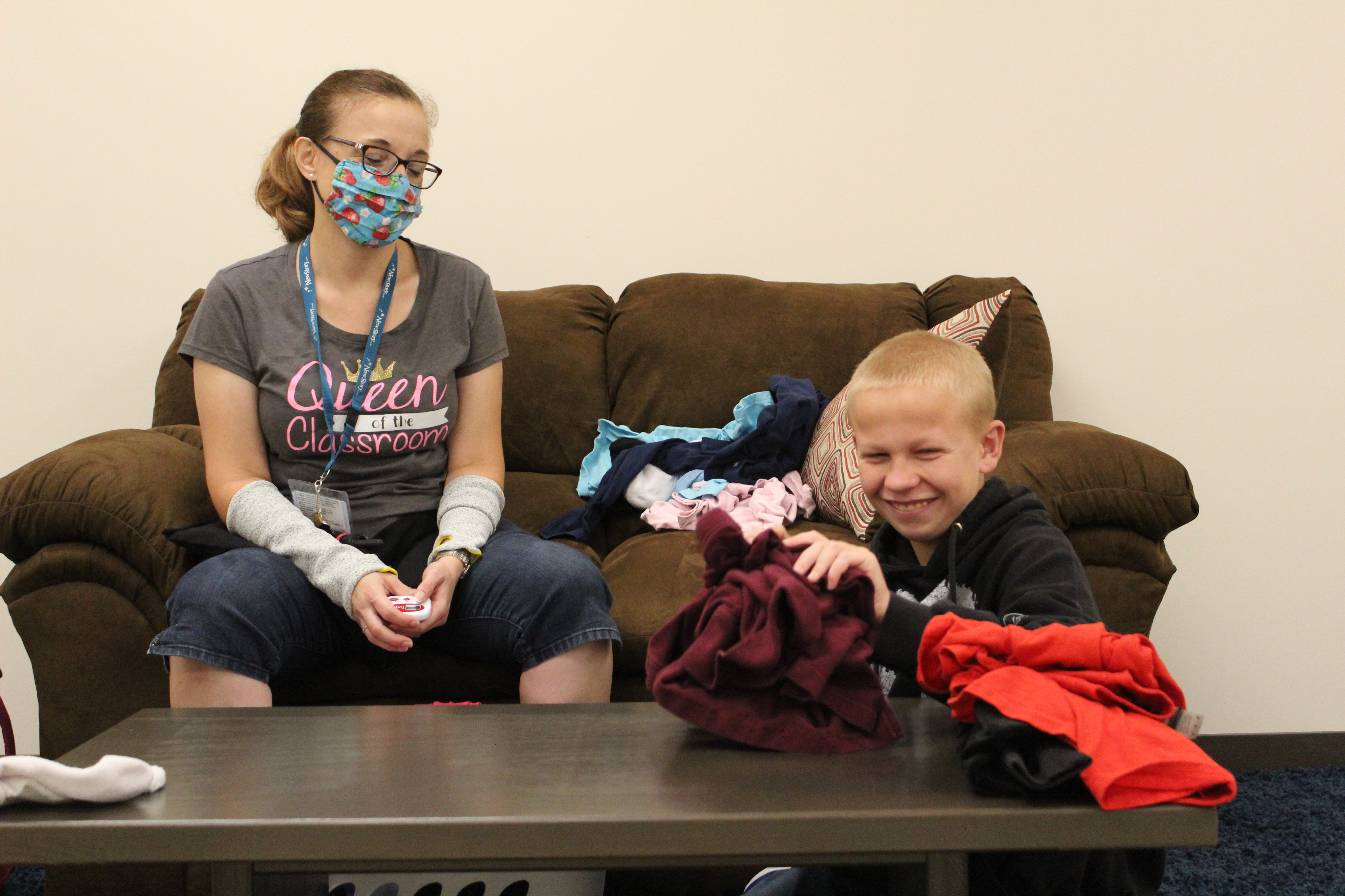 Special Education Student folds clothes in the Life Skills apartment. His Teacher, sitting on the couch, watches. The teacher is wearing a mask. 