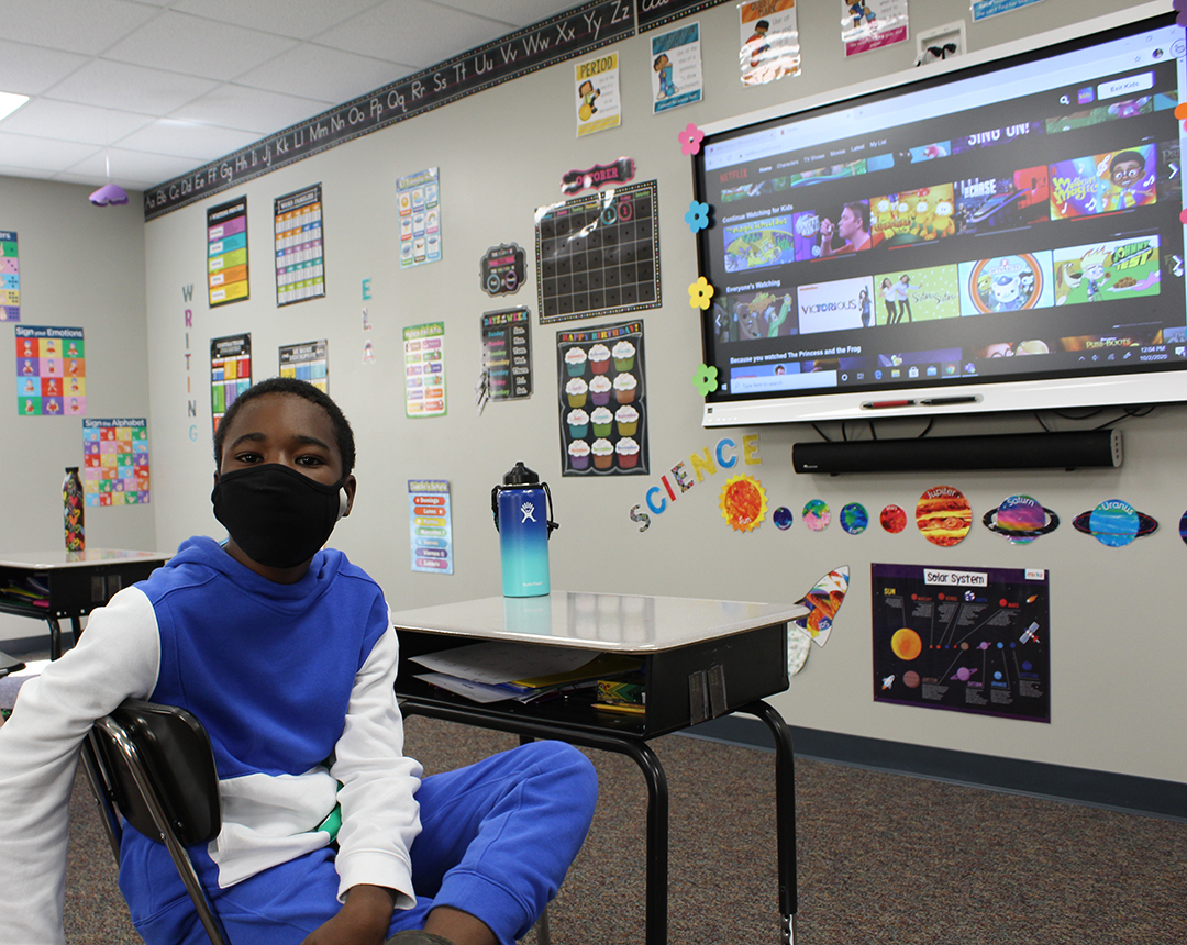 Special Education Student with mask sits at his front row desk in front of his classroom's smartboard, which is showing various kid's videos for break time music.