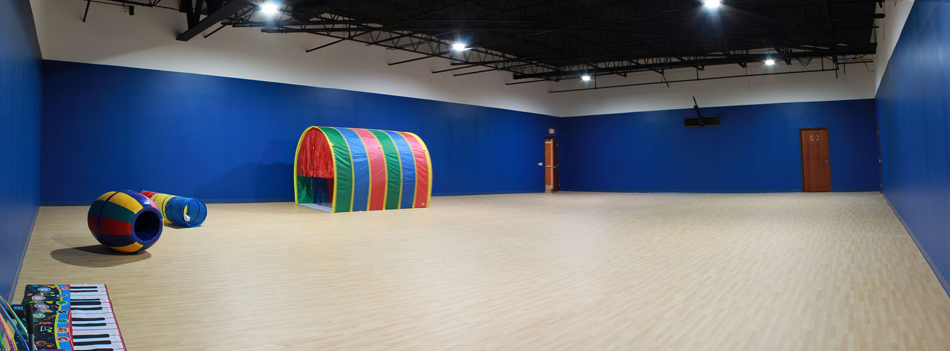 Gymnasium Bright but deep blue walls, high, lofty ceiling with exposed piping, light wood floor, bright toys scattered.